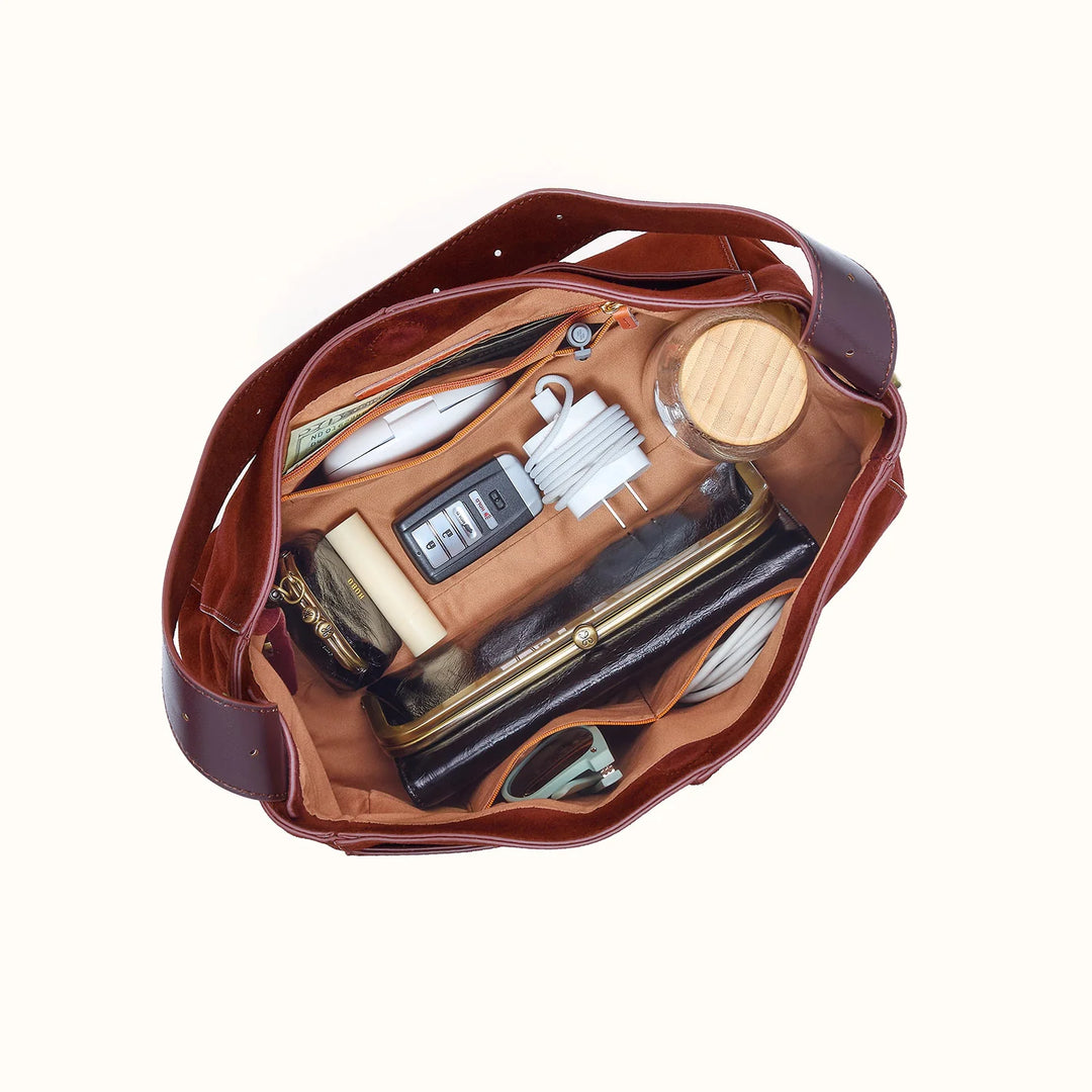 An overhead view of an open brown leather bag filled with various travel accessories, including a wooden compact, wallet, sunglasses, and other small items, set against a plain off-white background.